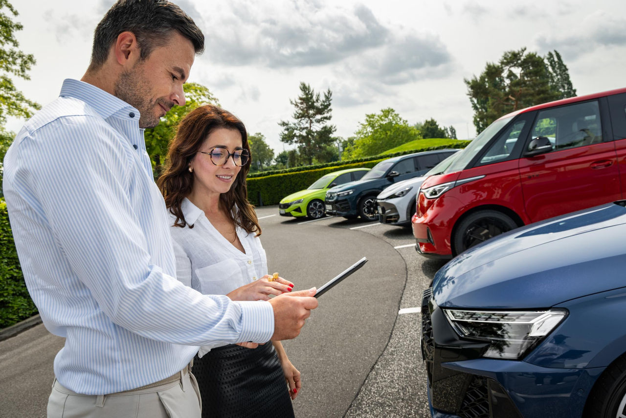Two people standing in a parking lot, looking at a tablet. Several cars, including a red van, white car, and blue car, are parked nearby. Trees and greenery are visible in the background under a cloudy sky.